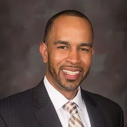 Professional studio portrait of Patrick Hairston. He is smiling and wearing a dark suit jacket, white shirt and striped tie.
