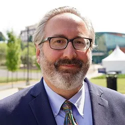 Photo of smiling man with glasses and a beard. He's wearing a light-colored shirt, dark tie and dark suit jacket.
