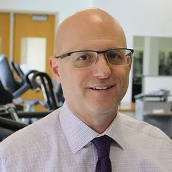 A smiling man with glasses with fitness equipment in the background. He is wearing a light-colored shirt and dark tie.